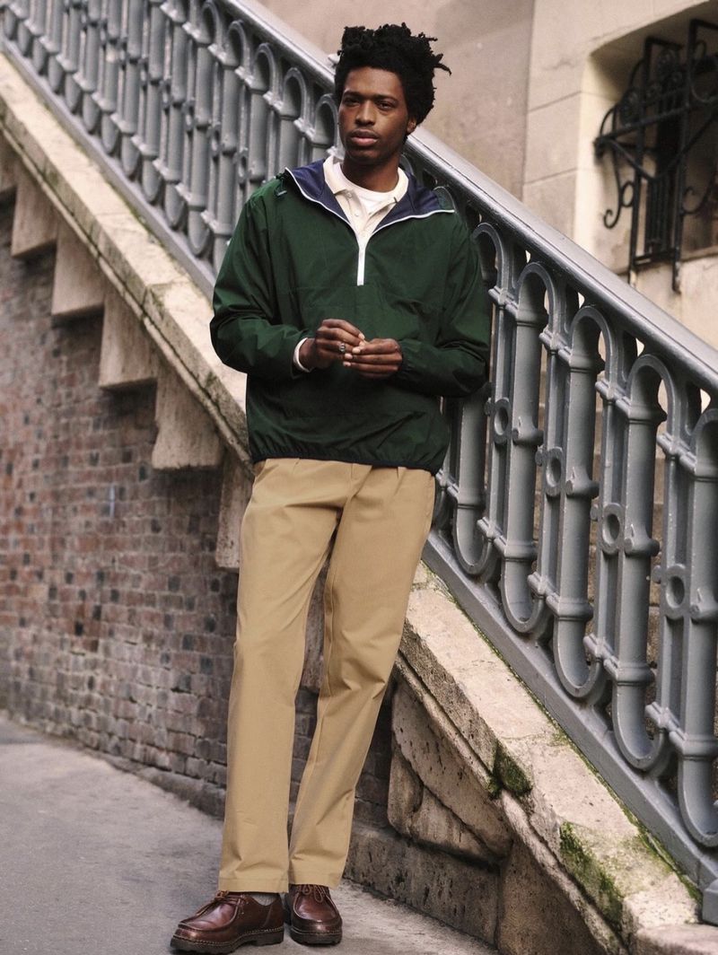 Full body shot of a Black male model wearing a dark green hooded jacket and khaki pants, posing on stone steps next to an iron railing.