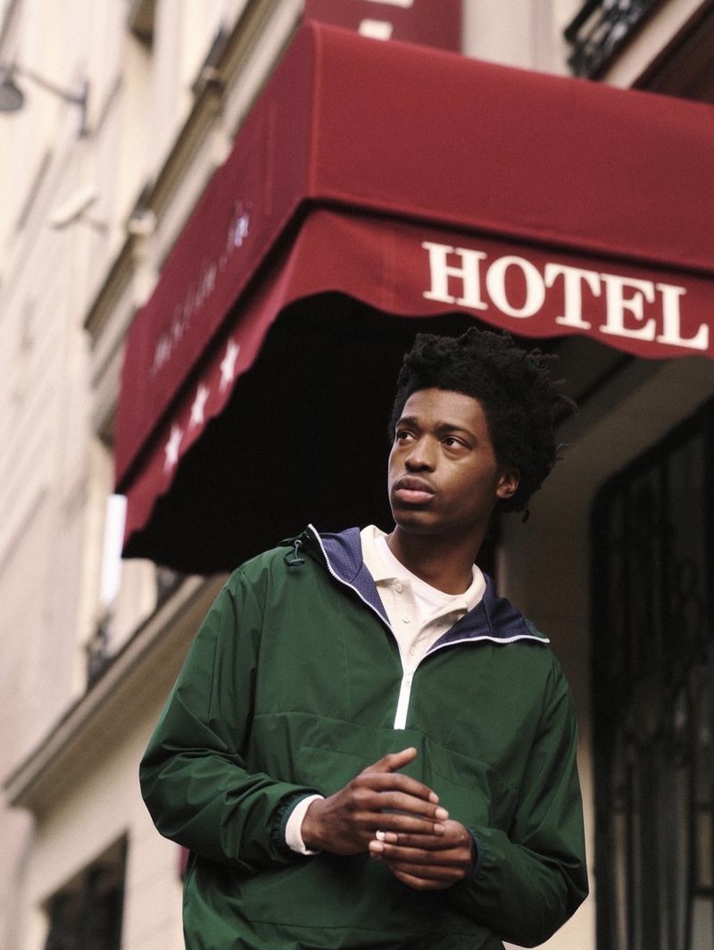 An eye-level portrait of a young adult man standing outdoors next to a hotel with a maroon awning and dark green windbreaker.