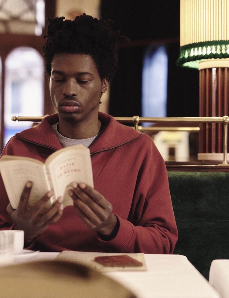 A young Black adult reads a book at a table, wearing a red zip-up sweatshirt. Indoor shot with soft lighting. Neutral expression.