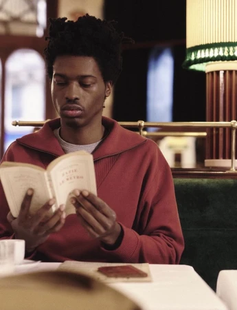 A young Black adult reads a book at a table, wearing a red zip-up sweatshirt. Indoor shot with soft lighting. Neutral expression.