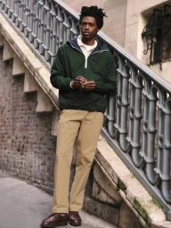 Black man wearing a dark green anorak and khaki trousers standing on outdoor stone steps next to an ornate metal railing.