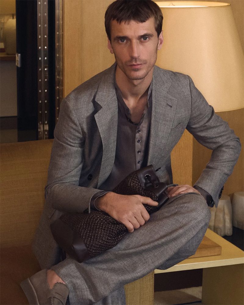 Portrait of a man in a grey suit seated indoors holding a brown textured bag, looking directly at the camera with neutral expression.