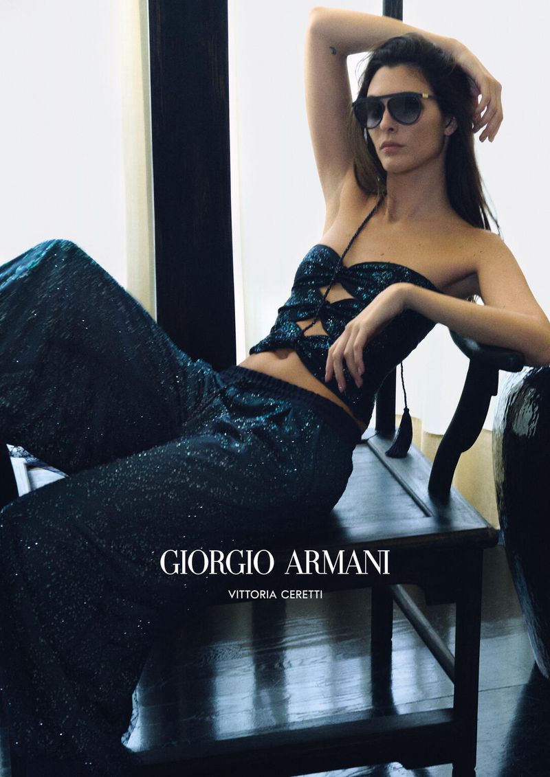 An adult woman poses in an indoor setting wearing sunglasses, a glittery halter top, and matching pants while sitting on a black chair.