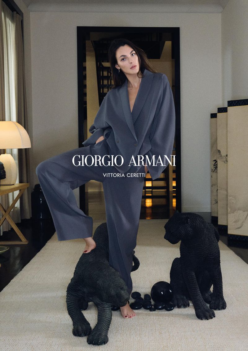 Elegant woman in gray loungewear poses indoors with decorative black panther sculptures on a light carpet in a high-fashion setting.