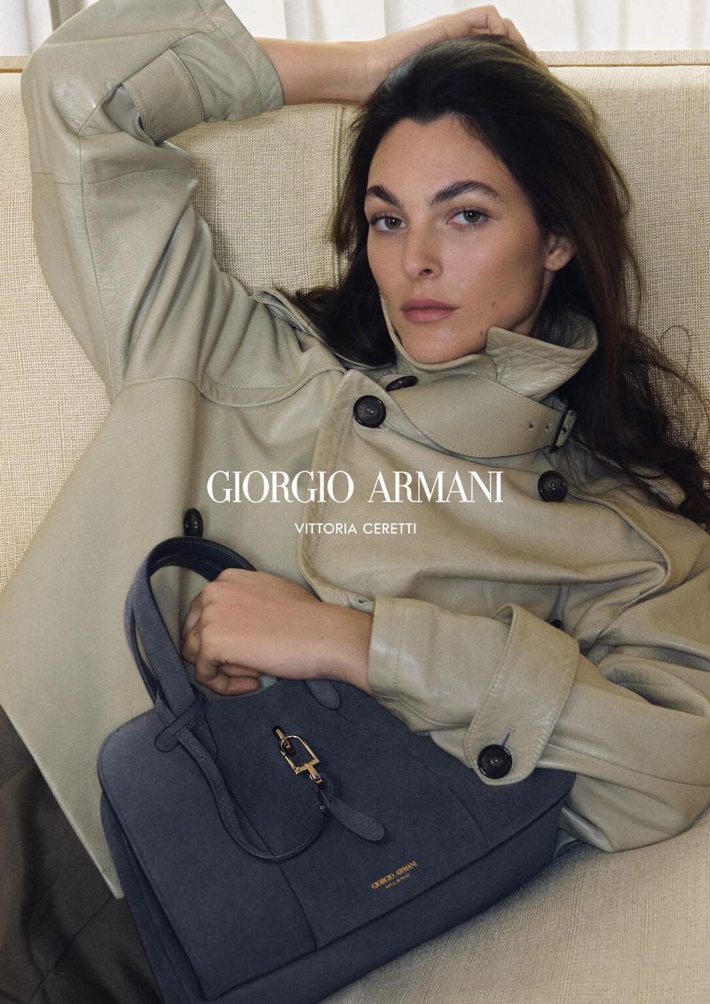 A brunette fashion model poses with a blue leather handbag in an indoor setting, wearing a light-green trench coat, advertising Giorgio Armani.