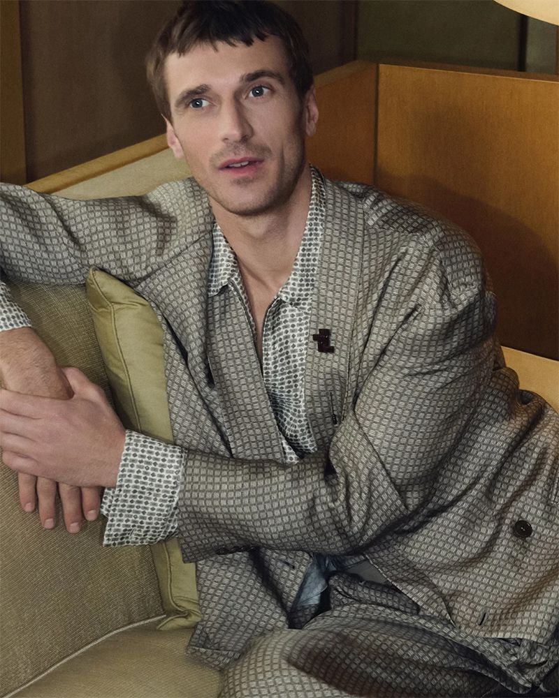 Indoor medium shot of a male model in patterned suit, seated on a beige couch looking slightly to the right, in a relaxed pose.