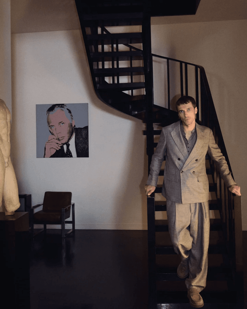 Adult man in a suit poses on dark stairs in front of a portrait and black staircase in an interior space with natural light.