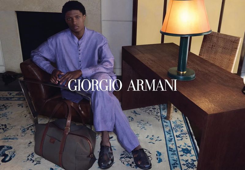 A dark-skinned male model is seated in a chair next to a dark-stained desk, in a relaxed pose, accessorized with a duffel bag and lit lamp.