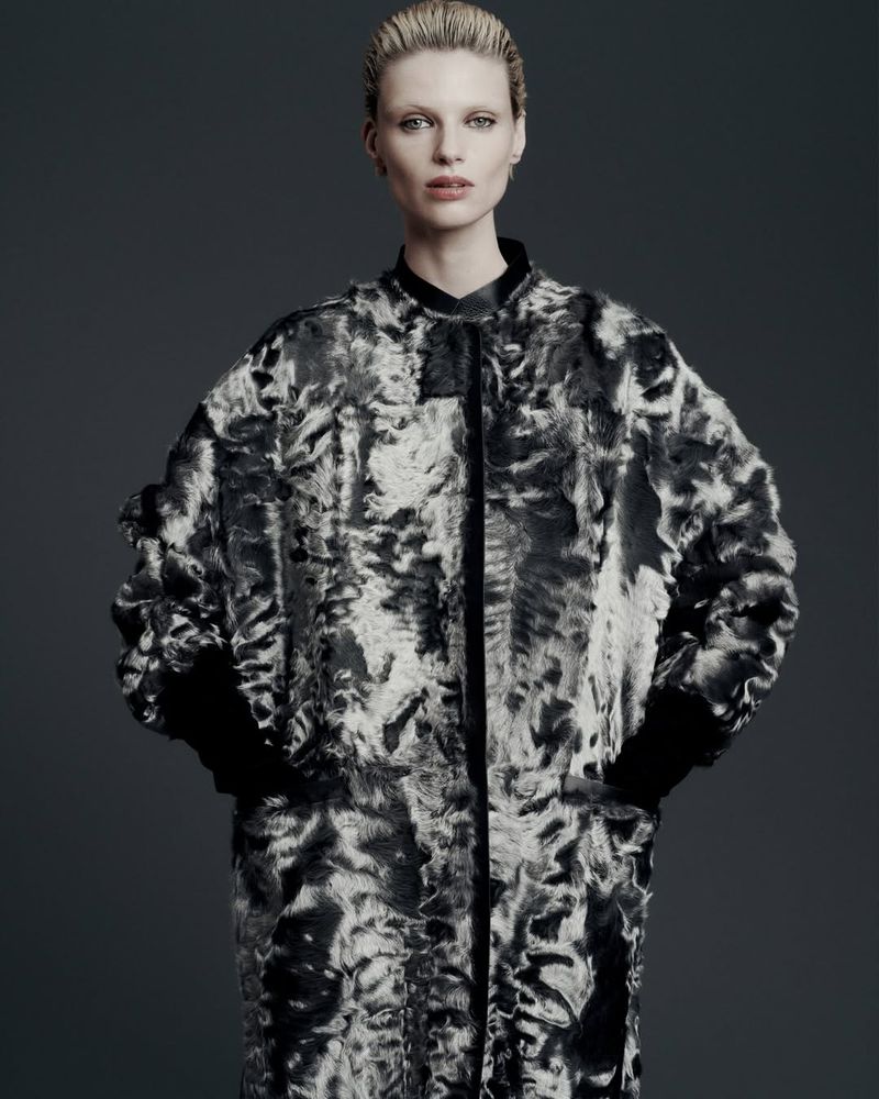 Woman in statement coat with gray and white abstract pattern, posing against a neutral gray backdrop in a fashion photography studio.