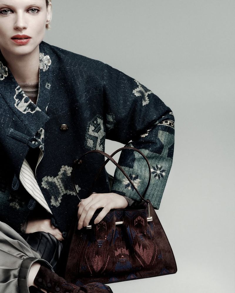 A fair-skinned model poses with a brown purse, wearing a patterned jacket and skirt, in a studio setting with neutral lighting.