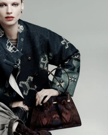 A fair-skinned model poses with a brown purse, wearing a patterned jacket and skirt, in a studio setting with neutral lighting.