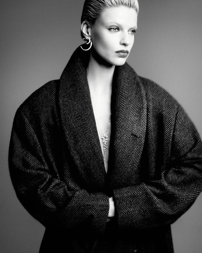Black and white portrait of an adult woman with a short hairstyle, wearing hoop earrings and a herringbone-patterned coat on a grey backdrop.