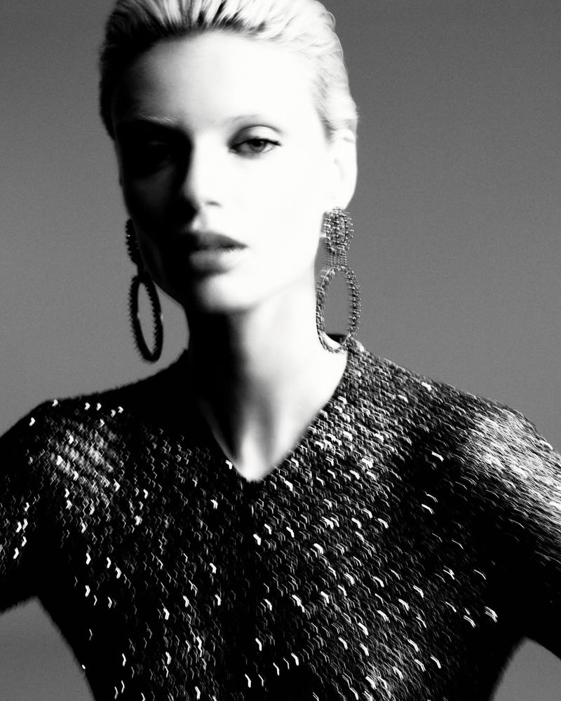 Black and white portrait of a blonde woman in a sequined top and statement earrings against a neutral gray backdrop in a photo studio.