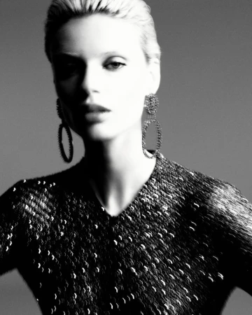 Black and white portrait of a blonde woman in a sequined top and statement earrings against a neutral gray backdrop in a photo studio.