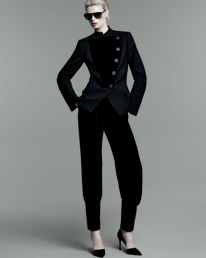 A full-length studio portrait of a stylish adult woman wearing a black velvet suit, black sunglasses, and high heels.