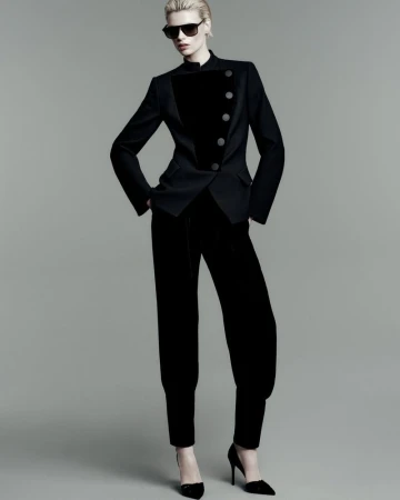 A full-length studio portrait of a stylish adult woman wearing a black velvet suit, black sunglasses, and high heels.