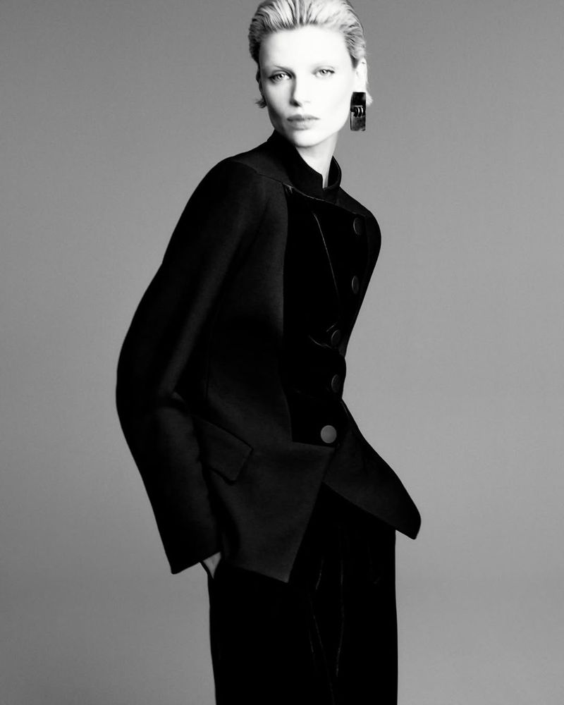Monochrome fashion portrait of a blonde model in a black velvet blazer and trousers against a neutral gray backdrop.