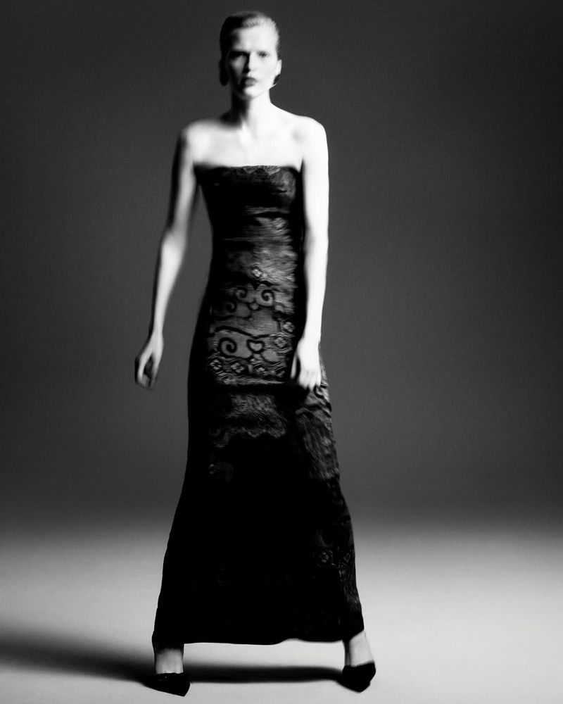 A slender woman posing in a strapless black lace gown and black high heels set against a dark gray backdrop in a studio setting.