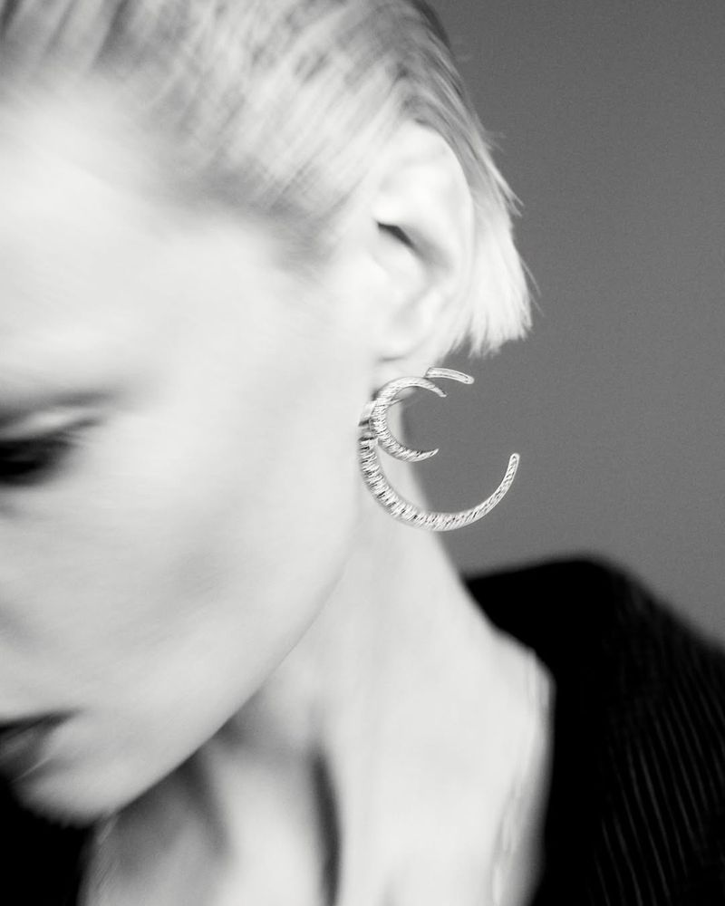 Close-up monochrome portrait of a model wearing distinctive crescent-shaped earrings against a grey background.