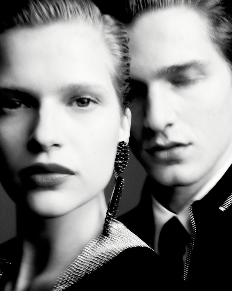 Monochromatic portrait of a blonde woman with long earrings, standing beside a dark-haired man in formal wear against a dark backdrop.