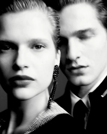 Monochromatic portrait of a blonde woman with long earrings, standing beside a dark-haired man in formal wear against a dark backdrop.