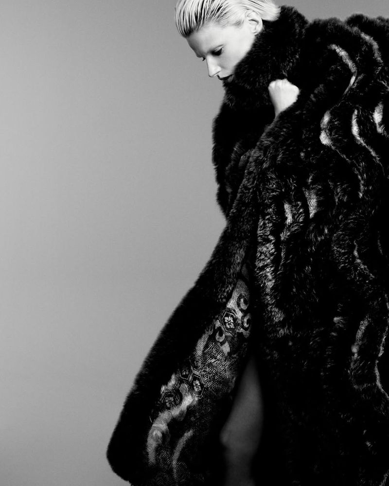 Black and white studio shot of a blonde woman in a dark fur coat, looking down. Fashion and portrait photography, posing in studio.