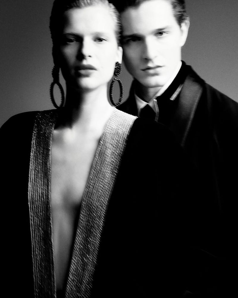 Black and white studio shot of a woman in open black and silver jacket and large earrings standing with a man in a black suit and tie.