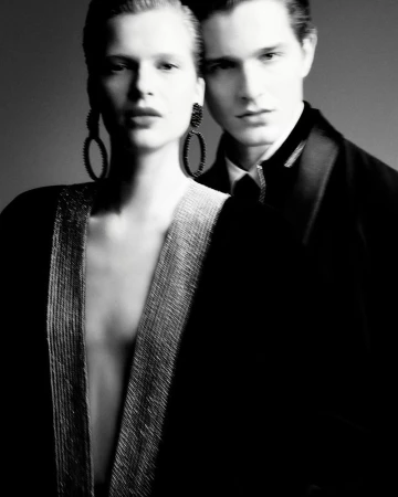 Black and white studio shot of a woman in open black and silver jacket and large earrings standing with a man in a black suit and tie.