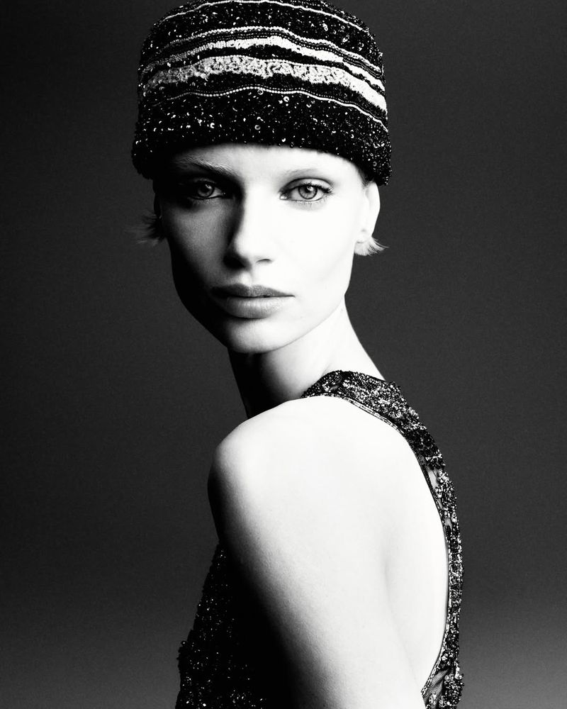 Black and white portrait of a woman with fair skin wearing a sequined hat and dress looking over her shoulder in a studio setting.
