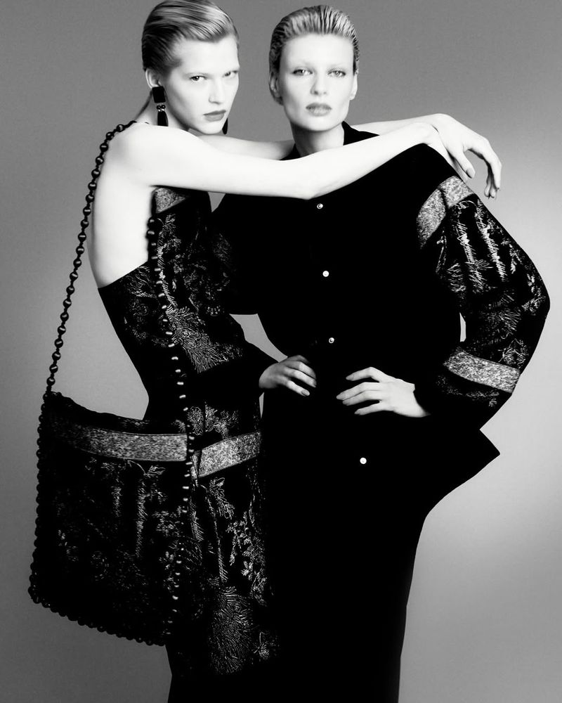 Monochrome fashion shot of two women in black outfits, one wearing a patterned dress and a large handbag, posing in a studio setting.