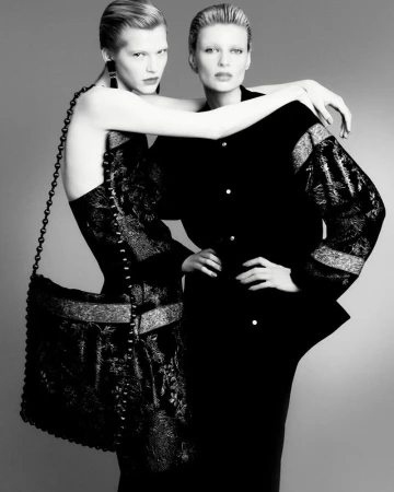 Monochrome fashion shot of two women in black outfits, one wearing a patterned dress and a large handbag, posing in a studio setting.