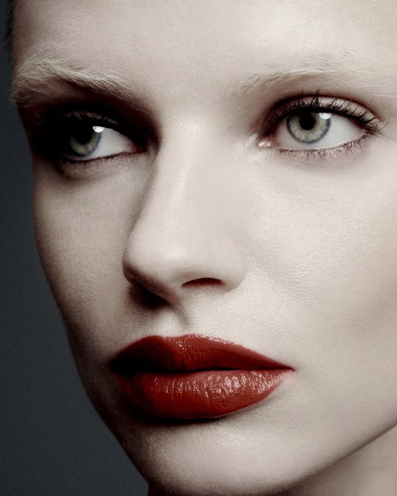 Close-up portrait of a woman with fair skin and pale blonde eyebrows, wearing glossy red lipstick and looking off to the side.