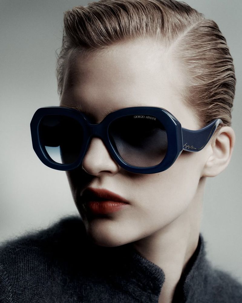 Close-up studio shot of a fair-skinned adult woman wearing dark framed sunglasses, red lipstick, and a grey jacket against a light grey backdrop.