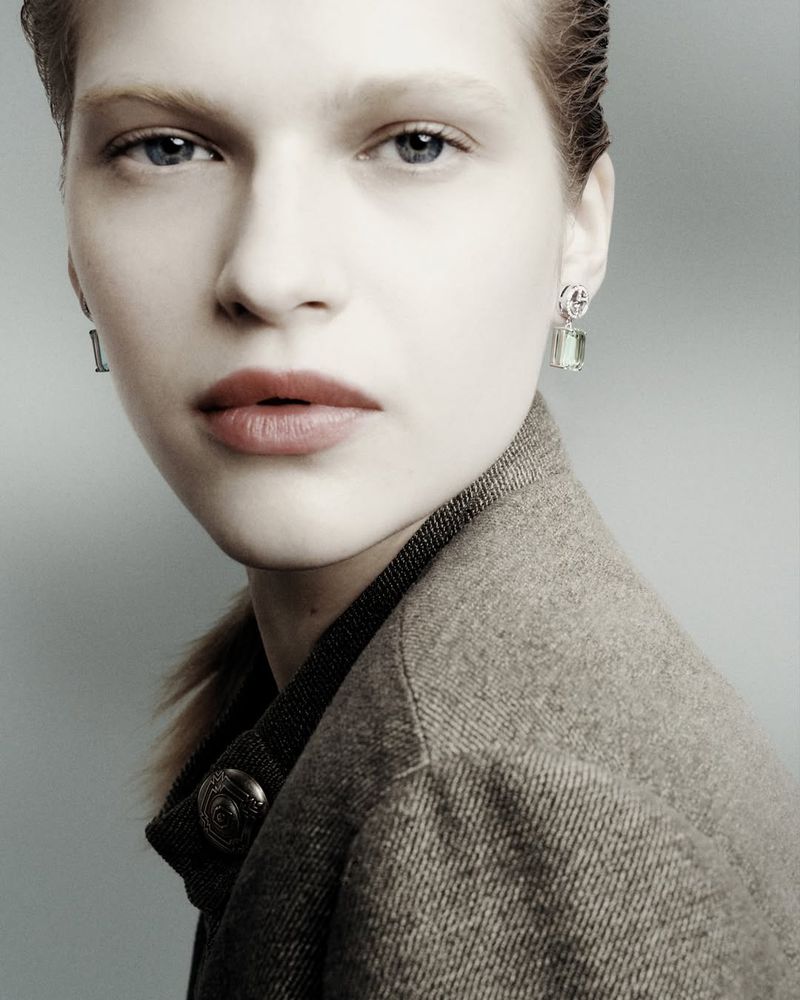 Headshot of a fair-skinned young adult woman in a grey wool jacket and statement earrings, set against a pale grey background. Neutral makeup.