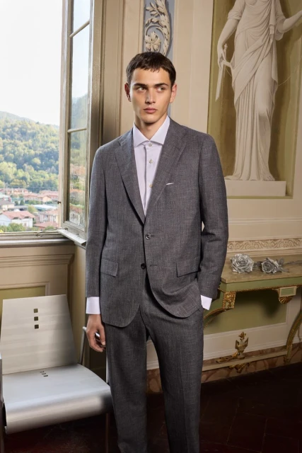 A young man in a gray suit and white shirt poses indoors near a window with a classical sculpture in the background.