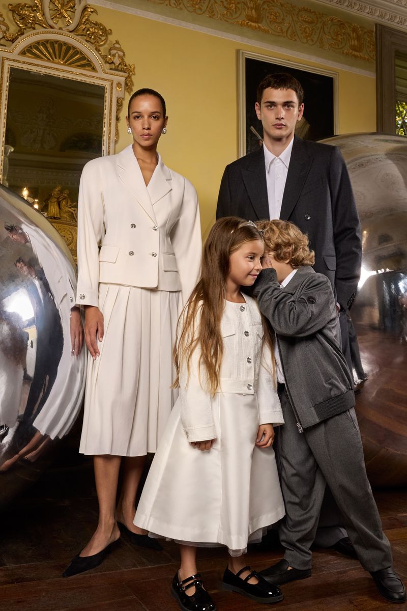 A family of four poses in formal wear indoors; the mother wears a cream suit, the father a black suit, and the children matching outfits.