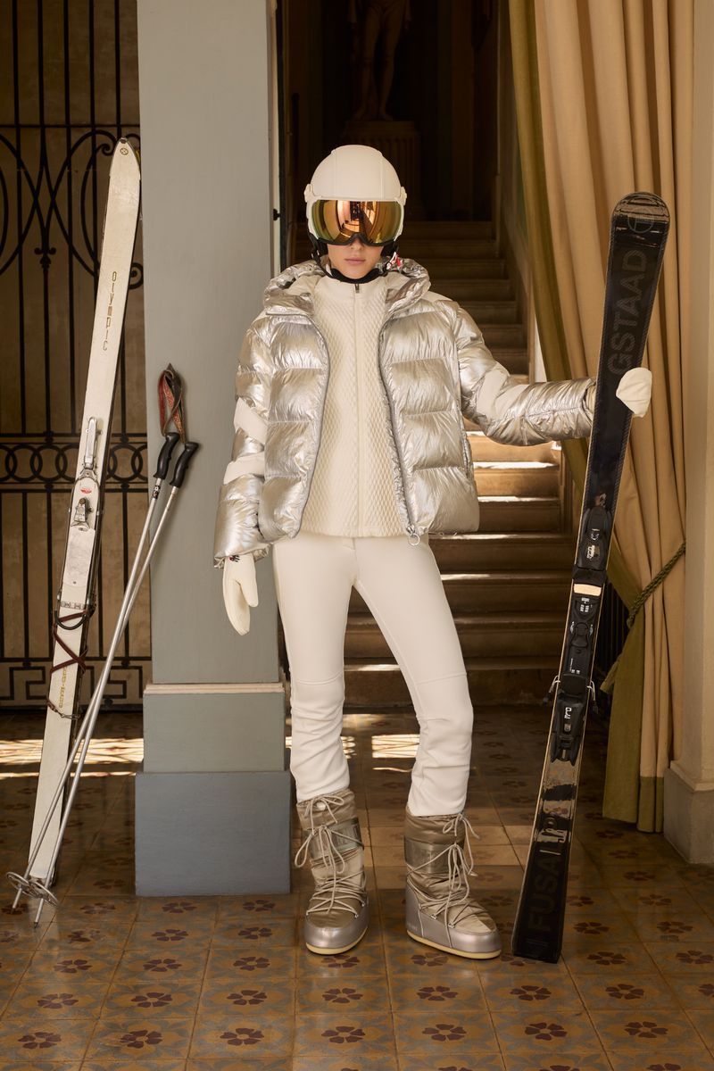 Model in a silver puffer jacket, white pants, and ski boots holding skis inside a building with a staircase and statue.