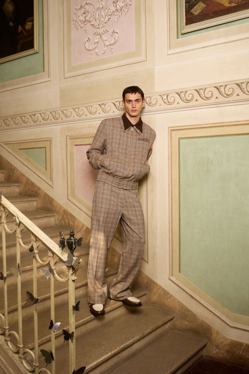 A young adult male in a patterned brown suit poses on stairs with butterfly-themed decorations in a brightly lit, indoor architectural space.