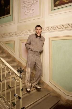 A young adult male in a patterned brown suit poses on stairs with butterfly-themed decorations in a brightly lit, indoor architectural space.