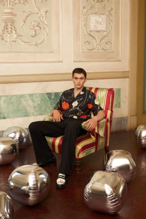 Man in floral shirt sits in striped chair surrounded by silver balloon spheres in ornate room with decorative wall panels and marble floor.