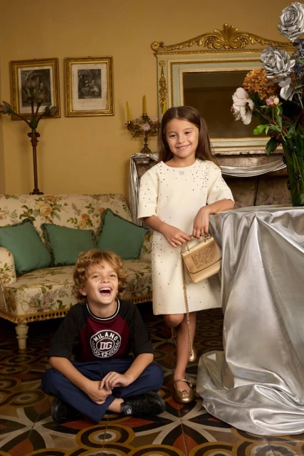 A smiling girl in a dress and a laughing boy in a t-shirt pose indoors in front of a couch, a decorative table, and wall decor.