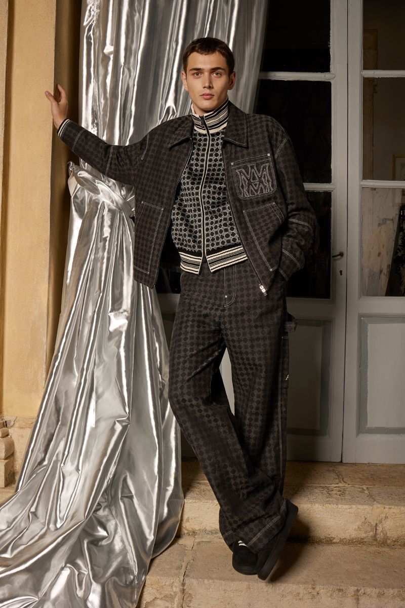 Man in grey patterned jacket and pants leans against a silver curtain next to a white doorway indoors, fashion editorial photography.