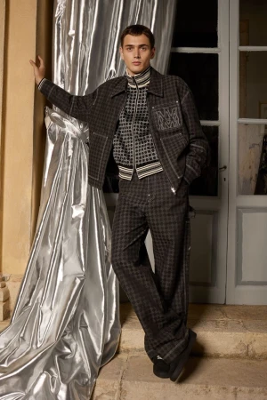 Man in grey patterned jacket and pants leans against a silver curtain next to a white doorway indoors, fashion editorial photography.