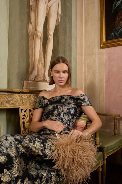 Elegant woman in a patterned off-the-shoulder gown sits holding a feathered purse with a statue and painting in the background.