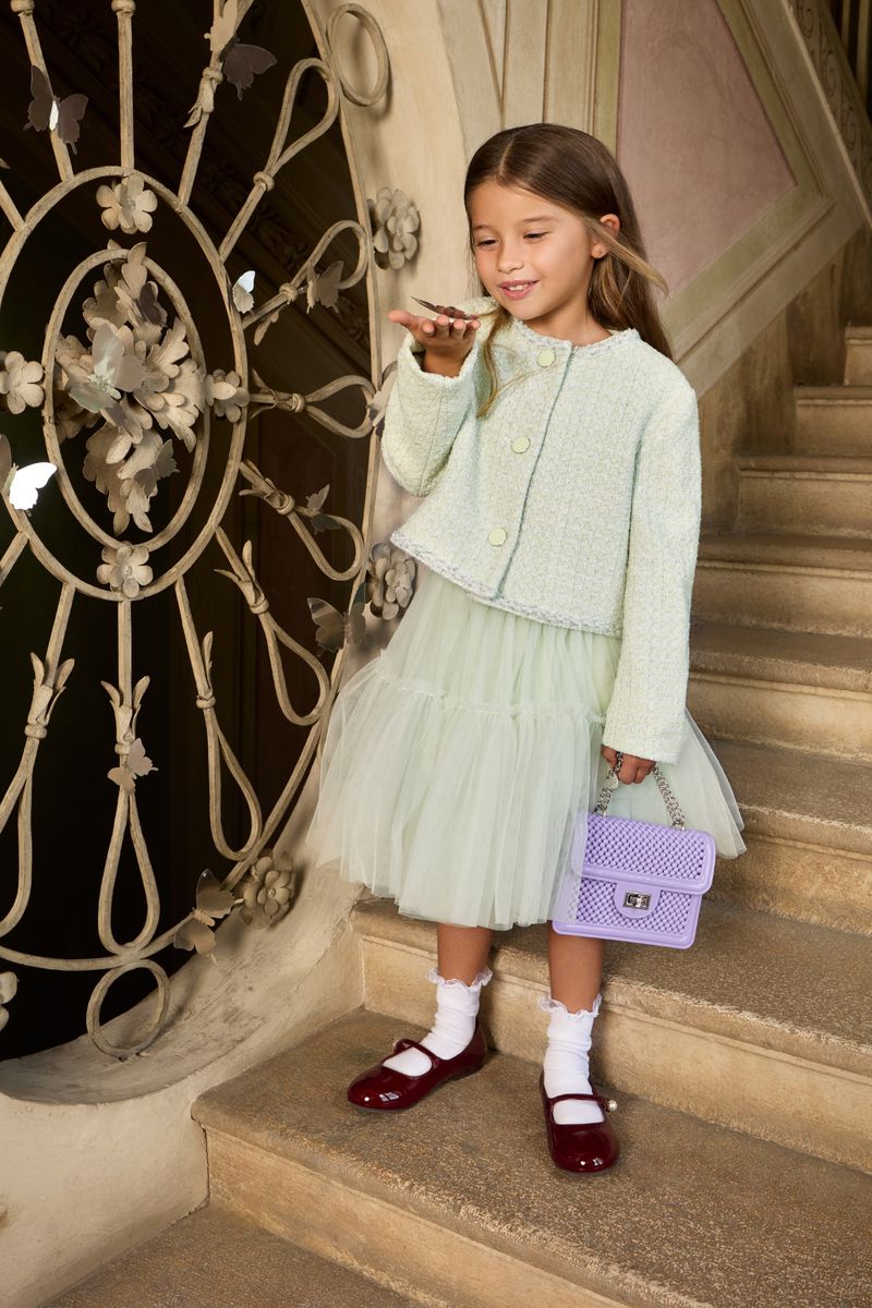 A young girl poses on stairs in light green jacket and tulle skirt, holding a small lavender purse with detailed wrought iron nearby.