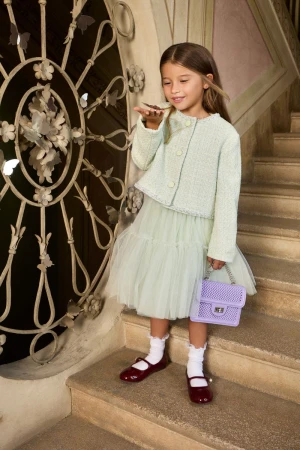A young girl poses on stairs in light green jacket and tulle skirt, holding a small lavender purse with detailed wrought iron nearby.