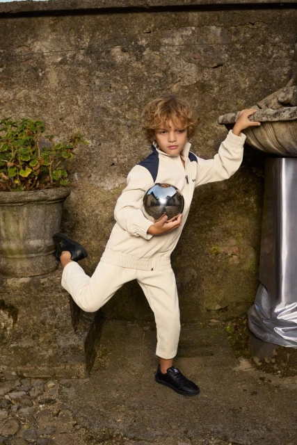 A young boy in a beige tracksuit poses outdoors with a shiny metal sphere, standing on a stone step near a weathered stone wall.