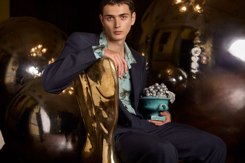 Man in suit poses with golden chair and ornate vase in dark studio setting, highlighting fashion and interior design elements.
