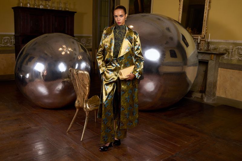 Adult female in a patterned gold coat and pants poses indoors near large metallic spheres and a gold chair, holding a tan purse.
