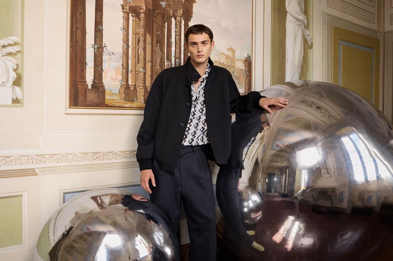 A male model poses indoors wearing a black jacket and patterned shirt, standing near large metallic orbs, in an ornate architectural setting.